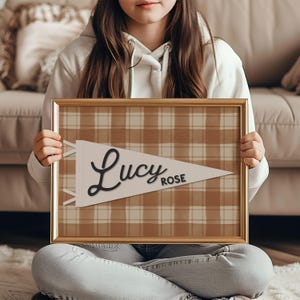 May include: A framed pennant-style print with a plaid background in shades of brown and cream. The pennant is white with the name "Lucy Rose" in elegant black script. The frame is a warm gold color.