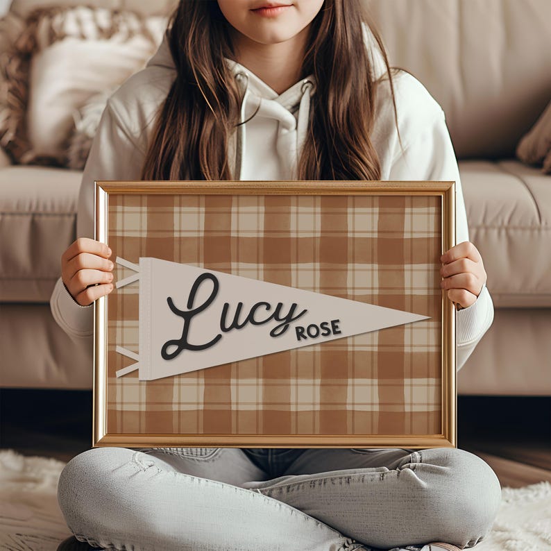 May include: A framed pennant-style print with a plaid background in shades of brown and cream. The pennant is white with the name "Lucy Rose" in elegant black script. The frame is a warm gold color.