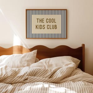 May include: A framed wall art piece with the text "THE COOL KIDS CLUB" in a brown font. The artwork is surrounded by blue vertical stripes and a brown frame. Below is a bed with striped bedding and a wooden headboard.