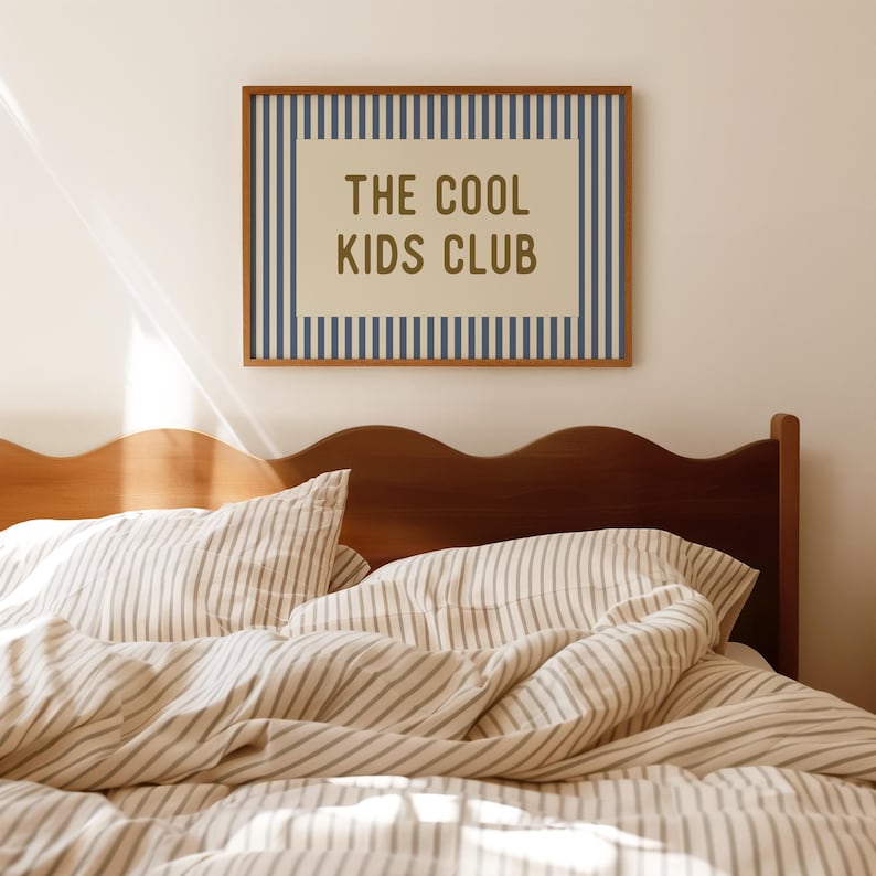 May include: A framed wall art piece with the text "THE COOL KIDS CLUB" in a brown font. The artwork is surrounded by blue vertical stripes and a brown frame. Below is a bed with striped bedding and a wooden headboard.