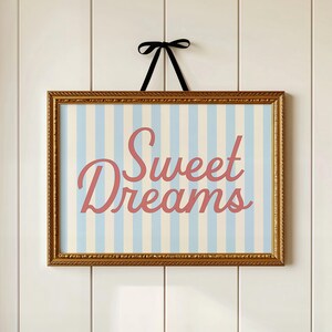 May include: A framed print with a gold-colored ornate frame. The print features a light blue and white striped background with the words "Sweet Dreams" in a cursive, coral-colored font. A black ribbon is attached to the top for hanging.