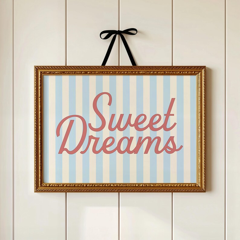 May include: A framed print with a gold-colored ornate frame. The print features a light blue and white striped background with the words "Sweet Dreams" in a cursive, coral-colored font. A black ribbon is attached to the top for hanging.