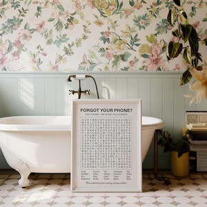 May include: A framed black and white word search puzzle with the title "Forgot Your Phone?" and the text "Don't worry, we've got you covered!"  The puzzle includes words related to bathroom items such as soap, towel, brush, and bath.  The puzzle is hanging on a white tiled wall in a bathroom.