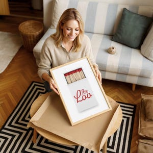 May include: A framed print featuring a white matchbox with red-tipped matches and the red text The Loo printed on the front. The artwork is encased in a light-colored wooden frame, showcasing a clean design with a focus on the matchbox illustration.