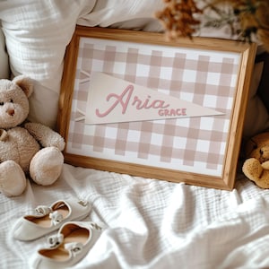 May include: A framed print featuring a tan and white gingham pattern. A pennant with the name "Aria" in pink script is centered on the print, with "Grace" below. A teddy bear and white baby shoes with bows are in the foreground.