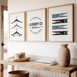 May include: Three framed prints with a laundry theme. The left print features a black silhouette of three hangers. The middle print has the text "The Johnson Family Laundry Co. Self Serve Open 7 Days A Week". The right print has a black and blue silhouette of four clothespins.