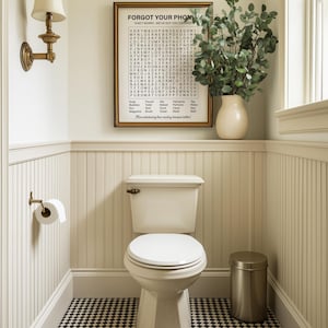 May include: A white toilet with a closed lid in a bathroom with white wainscoting and a black and white checkered floor. A framed print with the text "Forgot Your Phone? Don't worry, we've got you covered." is hanging on the wall above the toilet. A small potted plant is on the shelf above the toilet.