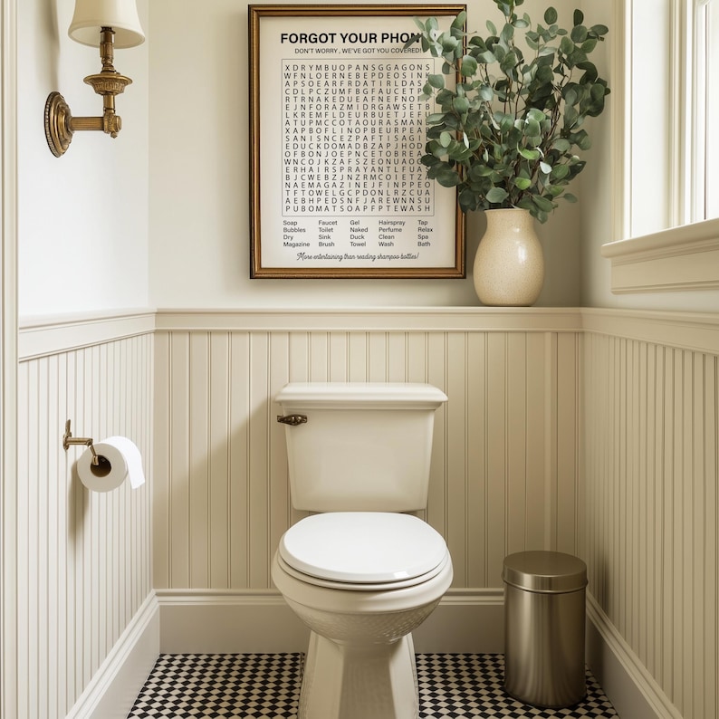 May include: A white toilet with a closed lid in a bathroom with white wainscoting and a black and white checkered floor. A framed print with the text "Forgot Your Phone? Don't worry, we've got you covered." is hanging on the wall above the toilet. A small potted plant is on the shelf above the toilet.