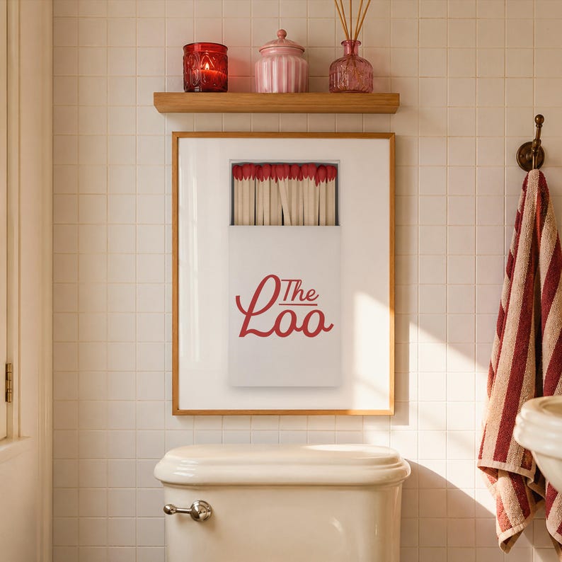 May include: A framed wall art print featuring a white matchbox with red-tipped matches and the red text The Loo. The print is displayed above a white toilet in a tiled bathroom, with a wooden shelf holding a red candle, a pink jar, and a reed diffuser.