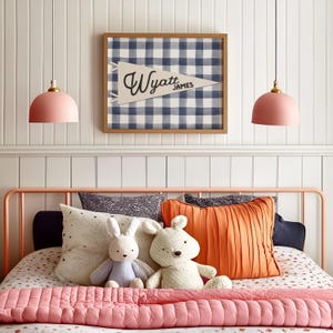 May include: A bedroom scene featuring a bed with a pink metal frame, a pink quilted blanket, and decorative pillows. Two plush bunny toys sit on the bed. Above the bed is a framed pennant with the name "Wyatt James" and a blue and white checkered pattern.