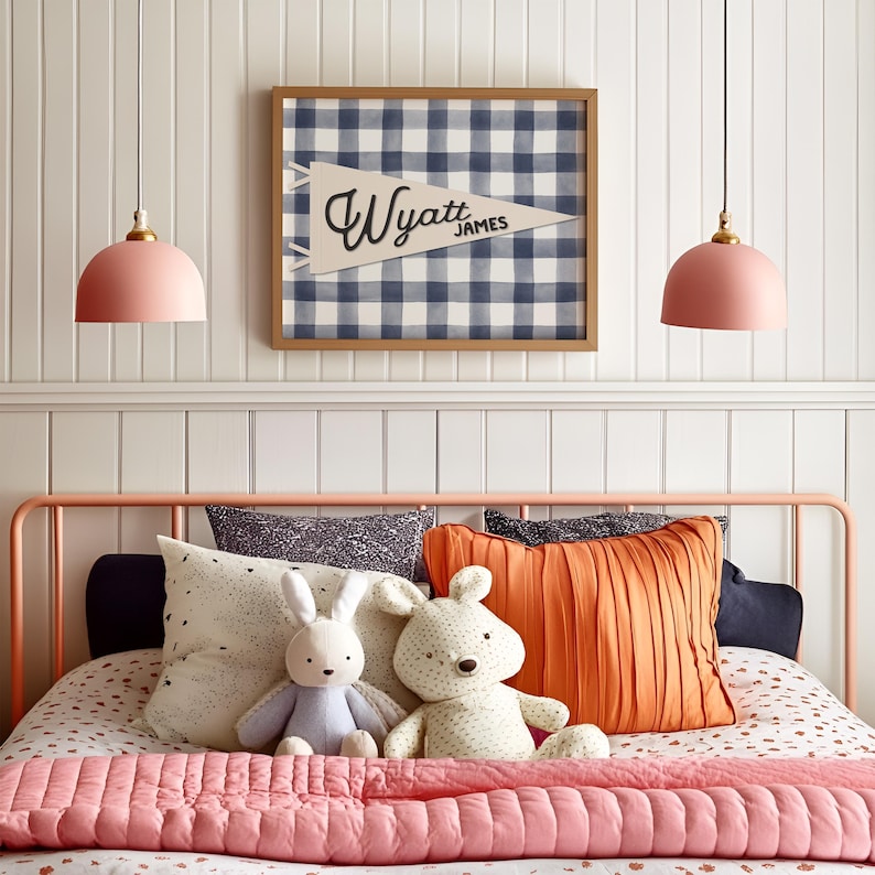 May include: A bedroom scene featuring a bed with a pink metal frame, a pink quilted blanket, and decorative pillows. Two plush bunny toys sit on the bed. Above the bed is a framed pennant with the name "Wyatt James" and a blue and white checkered pattern.