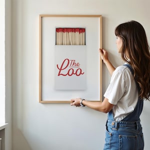 May include: A framed wall art print featuring a white matchbox with red match heads and the red cursive text The Loo. The print is encased in a light-colored wooden frame, displaying a graphic illustration of a matchbox filled with wooden matches.