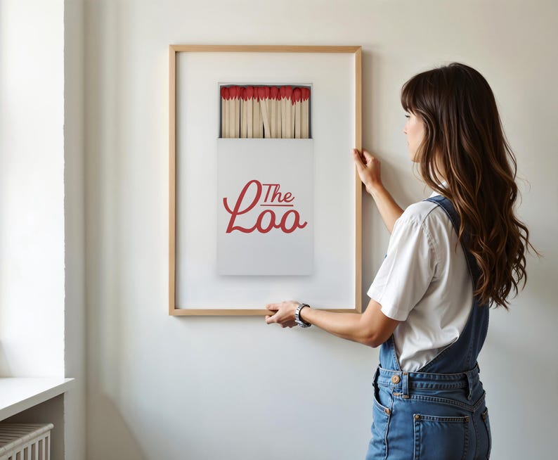 May include: A framed wall art print featuring a white matchbox with red match heads and the red cursive text The Loo. The print is encased in a light-colored wooden frame, displaying a graphic illustration of a matchbox filled with wooden matches.