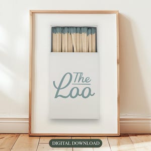 May include: A framed print of a white box with a blue and white design. The box contains a full pack of matches with blue tips. The text on the box reads "The Loo".