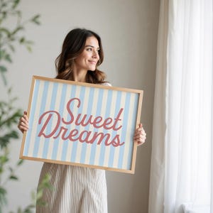 May include: A framed sign with a light blue and white striped background and the words "Sweet Dreams" in a cursive, red font. The frame is made of light-colored wood. The sign is held by a person.