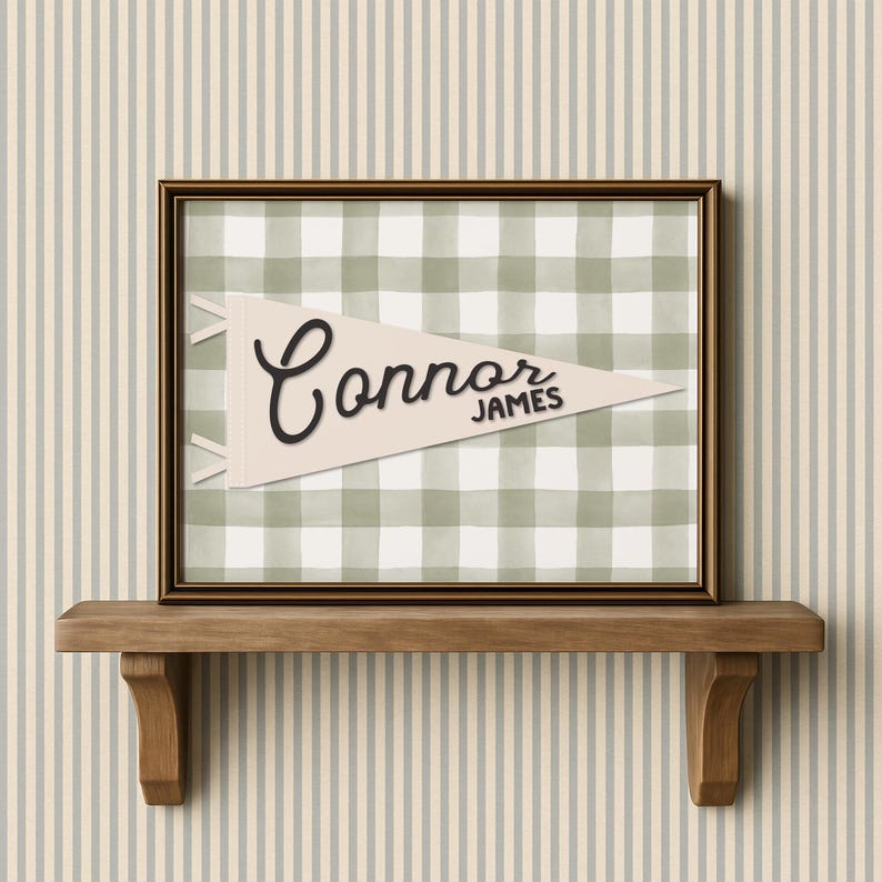 May include: A framed print featuring a pennant design with the name "Connor James" in black script. The pennant is set against a green and white gingham pattern. The frame is dark brown, and the print is displayed on a wooden shelf.