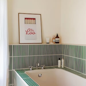 May include: A framed wall print featuring a white matchbox with red match heads and the red text The Loo. The print is displayed above a bathtub surrounded by dark green vertical tiles, with various bottles placed on the tiled ledge nearby.