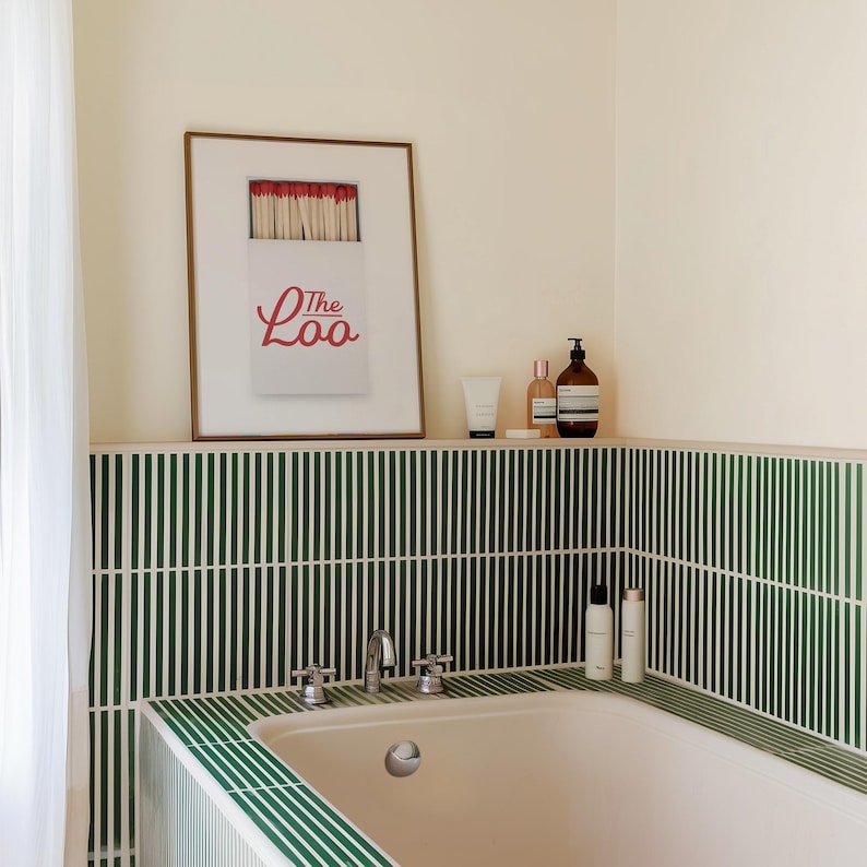 May include: A framed wall print featuring a white matchbox with red match heads and the red text The Loo. The print is displayed above a bathtub surrounded by dark green vertical tiles, with various bottles placed on the tiled ledge nearby.