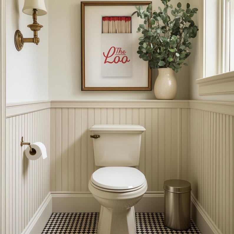May include: A bathroom interior featuring a white toilet with a closed lid, a wall-mounted toilet paper holder, a metallic trash can, and a framed print depicting a matchbox with the red text The Loo, accented by a brass wall sconce and greenery.