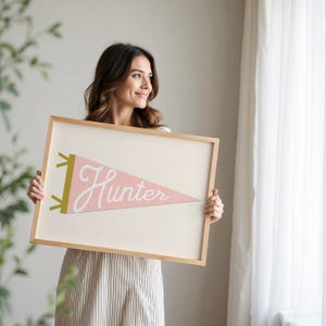 May include: A framed pink and white pennant with the word "Hunter" in white script. The pennant is in a wooden frame.