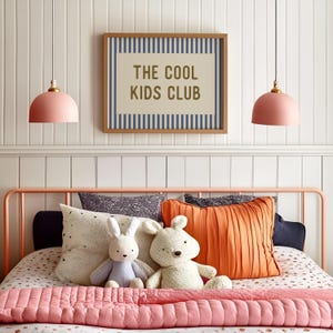 May include: A bedroom scene featuring a bed with a pink metal frame, adorned with pillows, stuffed animals, and a pink quilted blanket. Above the bed, a framed sign reads "THE COOL KIDS CLUB." Two pink pendant lights hang from the ceiling.