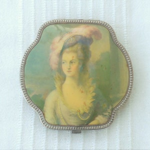 Könnte beinhalten: Ein Vintage-Kompaktspiegel mit silberfarbenem Rahmen und Kleeblattform. Das Bild auf dem Kompaktspiegel zeigt ein Porträt einer Frau in historischer Kleidung mit Federhut. Der Hintergrund ist eine weiche, pastellfarbene Landschaft.