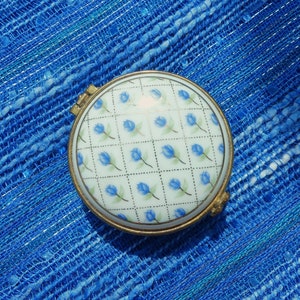 May include: A small, round, white porcelain trinket box with a hinged lid. The box is decorated with a repeating pattern of blue flowers on a white background. The box is surrounded by a gold metal frame.