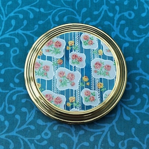 Puede incluir: Un espejo compacto redondo de color dorado con un diseño floral azul y blanco. El diseño presenta rosas rosas y hojas blancas sobre un fondo azul con rayas blancas.