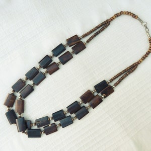 以下が含まれることがあります： 長方形のビーズが付いた茶色の木製ビーズネックレス。ネックレスは2連で、銀色の留め具が付いています。