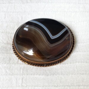 May include: A brown agate gemstone brooch with a copper-toned metal setting. The agate has a swirling pattern of brown, black, and white.