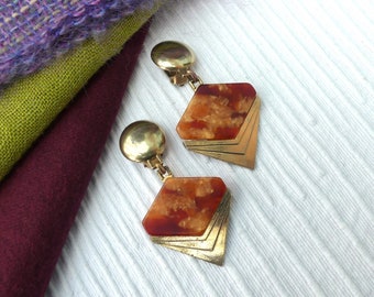 May include: A pair of gold tone clip-on earrings with a geometric design. The earrings feature a large, brown and orange, marbled, diamond-shaped stone.