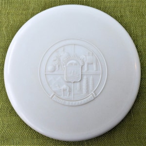 Könnte beinhalten: Ein weißer runder Plastikdeckel mit einem erhabenen Prägemuster eines Wappens mit einem Schiff, einem Kamel, einer Palme und anderen Symbolen. Der Text "India Septentrionalis" ist um das Muster herum geprägt.