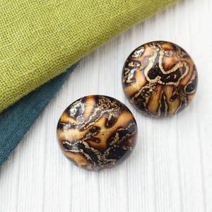 May include: Two round, decorative buttons with a brown and gold marbled design. The buttons are on a white surface with green and blue fabric in the background. The buttons have a glossy finish.