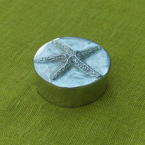 May include: A small, round, silver-colored trinket box with a starfish design on the lid. The starfish is raised and textured, set against a sandy background. The box sits on a green, textured surface.