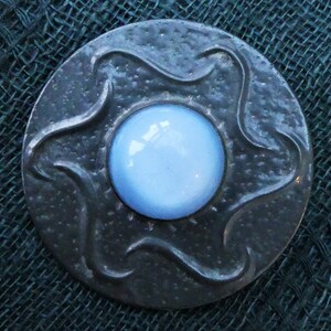 May include: A silver-toned brooch with a blue glass center. The brooch has a textured, wavy design around the edge.