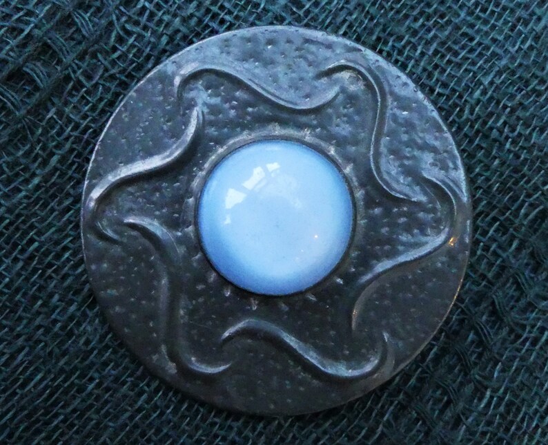 May include: A silver-toned brooch with a blue glass center. The brooch has a textured, wavy design around the edge.