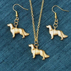 May include: A gold-colored jewelry set featuring a dog design. The set includes a necklace pendant and two earrings. The dog pendants are detailed and suspended from gold chains and earring hooks. The jewelry is displayed on a textured teal background.