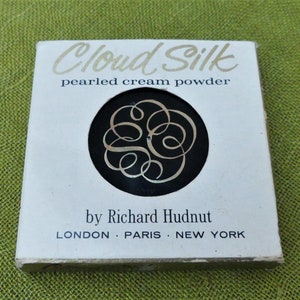 May include: A white box with gold lettering that reads "Cloud Silk pearled cream powder by Richard Hudnut LONDON PARIS NEW YORK". The box has a black circle with a gold swirl design.