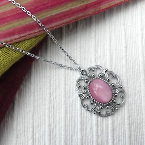 Puede incluir: Un collar con colgante plateado con una piedra ovalada rosa claro engastada en un marco calado ornamentado. La cadena del collar es delicada y el colgante descansa sobre una superficie blanca texturizada. Una bufanda colorida está en el fondo.