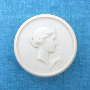 May include: A white ceramic round trinket box with a raised design of a woman's profile and a deer. The woman has a crescent moon above her head.