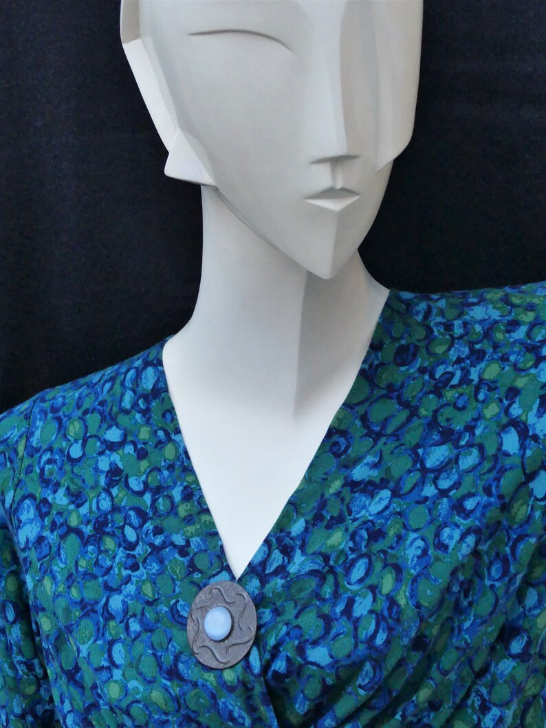 May include: A blue and green patterned blouse with a silver and blue brooch. The brooch has a round, blue stone in the center.
