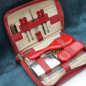 Op de afbeelding: Een rode leren reistas met een ritssluiting. De tas is open en bevat verschillende verzorgingsproducten, waaronder een haarborstel, een kam, een nagelvijl en een klein flesje.