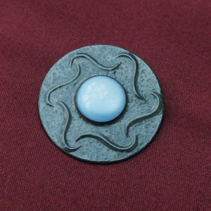 May include: A round, dark gray metal brooch with a light blue glass center. The metal has a textured, wavy design.