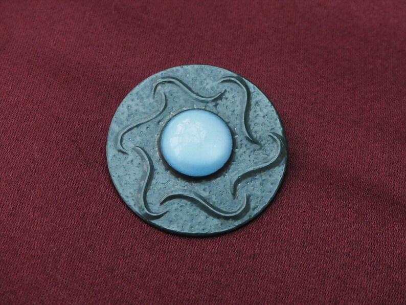 May include: A round, dark gray metal brooch with a light blue glass center. The metal has a textured, wavy design.