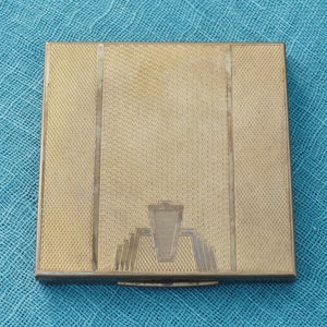 May include: A gold-colored metal compact with a geometric design and a textured surface.