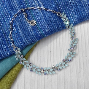 Puede incluir: Un collar plateado con diseño de hojas, con hojas de esmalte azul pálido y detalles de cristal transparente. El collar se encuentra sobre un fondo de tela azul y verde.