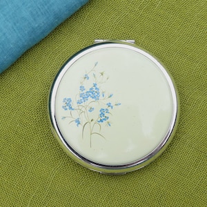 Op de afbeelding: Een zilveren compact spiegel met een witte achtergrond en een blauw bloemenontwerp. De spiegel heeft een zilveren rand en een bloemenontwerp aan de voorkant.
