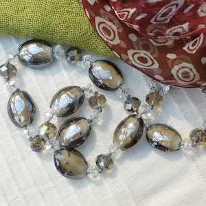 Puede incluir: Un collar de cuentas de vidrio plateado y gris con detalles de cristal transparente. El collar está sobre un fondo de tela blanca.