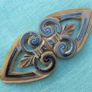 Puede incluir: Una hebilla decorativa marrón y negra con un diseño de espiral. La hebilla está hecha de un material plástico duro y tiene un acabado brillante.