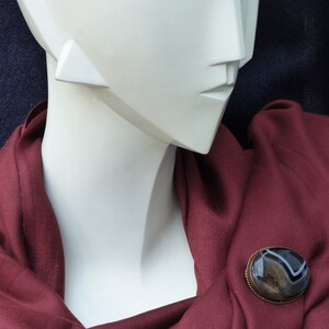 May include: A maroon scarf with a brown and white agate brooch. The brooch is oval shaped and has a silver setting.
